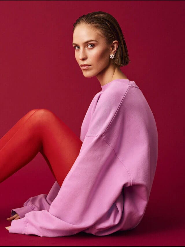 Lady with slicked back hair wears red tights and a pink jumper, with a red background
