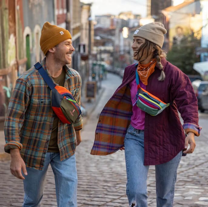 A man and woman wearing bright clothes, hats, and bags walk up a street laughing together