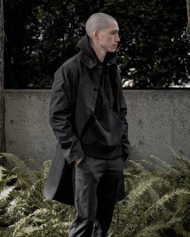 Bald man wearing all black; hoodie, trousers and trench coat, with trees and bushes in the background