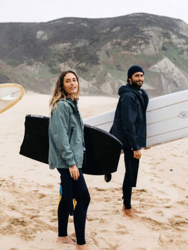A man and woman stand on the beach holding surfboards, wearing wet suits and coats