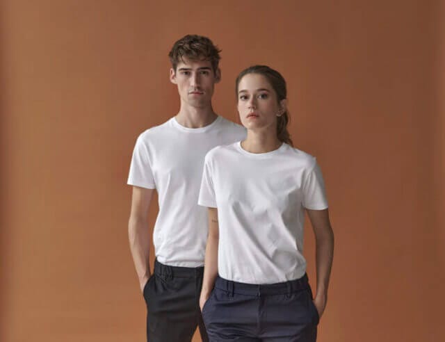 Man and woman standing side by side both wearing smart black trousers and white t-shirts with an orange background