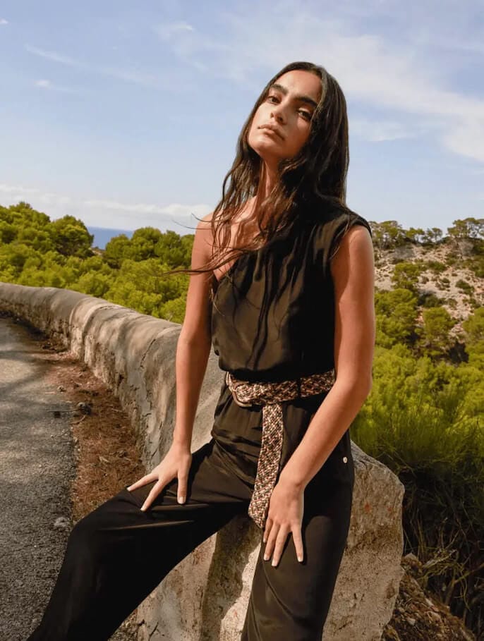 Woman sitting on a stone wall with sand and trees behind her wears a black jumpsuit with a brown belt