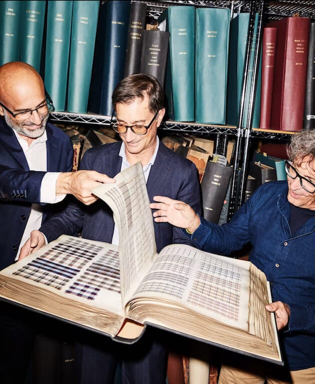 Three men wearing glasses look at patterns in a big book