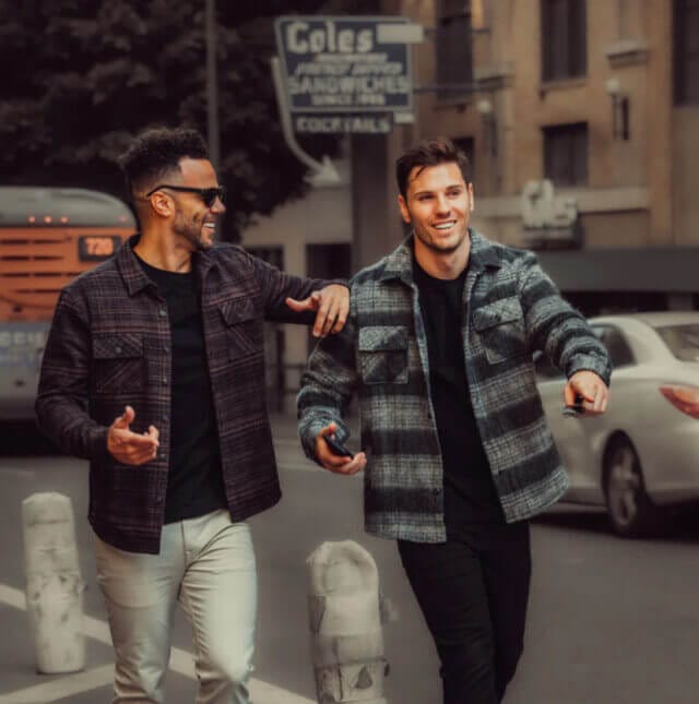 Two men wearing plaid jackets walk down the street together smiling