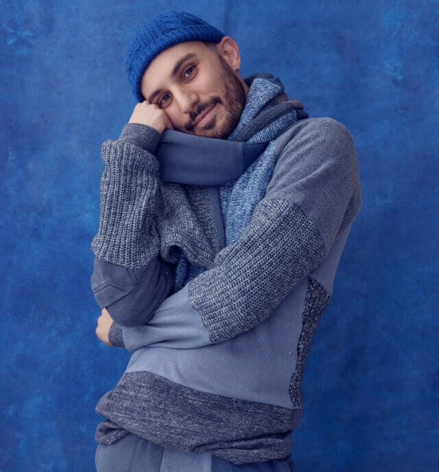 Person wearing blue knitted hat, jumper, and scarf stands with hand on chin