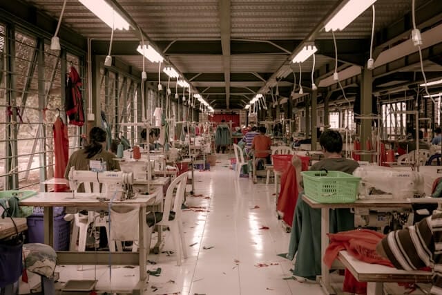 Inside a textile fabric, there are some desks with sewing machines, baskets, textile pieces on the floor and people working giving their backs to the viewer.