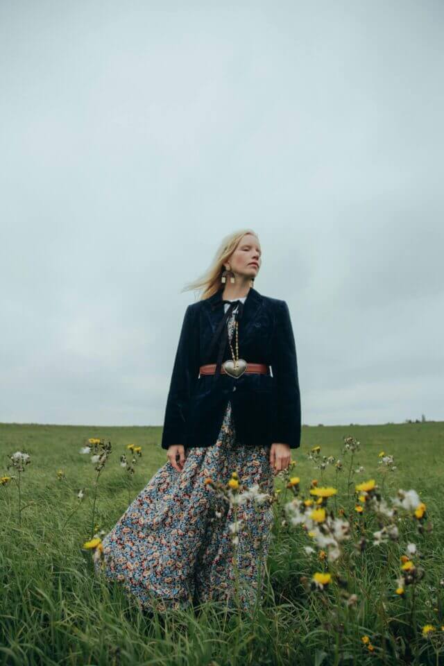 A blond woman stands in an open field with her eyes closed. She wears a hippy outfit: a velvet navy jacket, a tan waist belt and a flower dress.