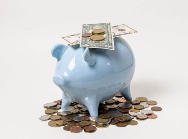 A little blue piggy bank stands on a cream background. It stands on coins and has some dollars and coins on the top.