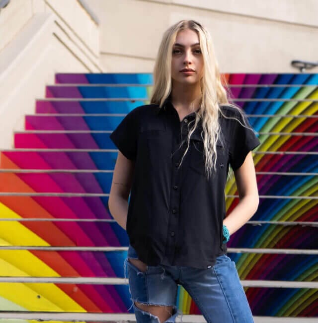 A woman stands in front of multicolor steps with a Switchback Shirt and a jeans.