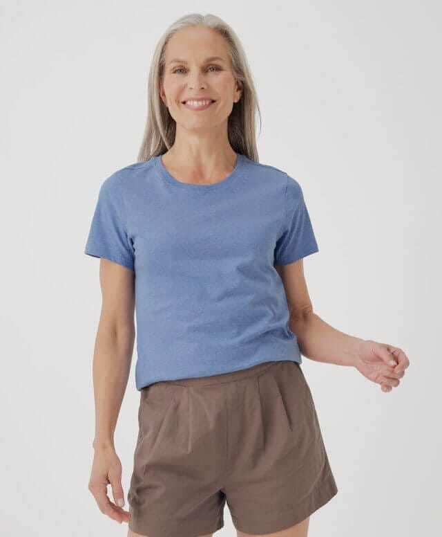 A woman stands on a white background with a brown short and a light blue t-shirt from Pact made with organic cotton in a fair trade factory.