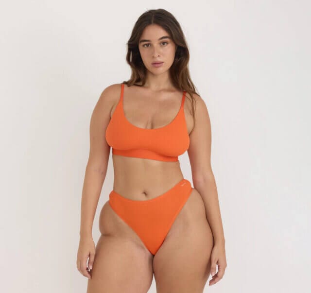 A tall woman with black hair wears a bright orange bra with a core rib thong made with organic cotton by Organic Basics.