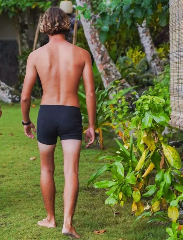Back view of a male model walking barefoot on grass, wearing black boxer briefs made from hemp and organic cotton, surrounded by tropical greenery.