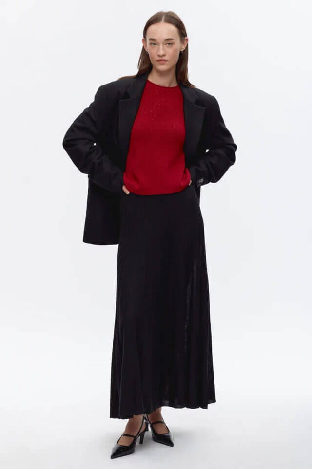 A woman standing against a white background wearing a red top, long black blazer and long black skirt.
