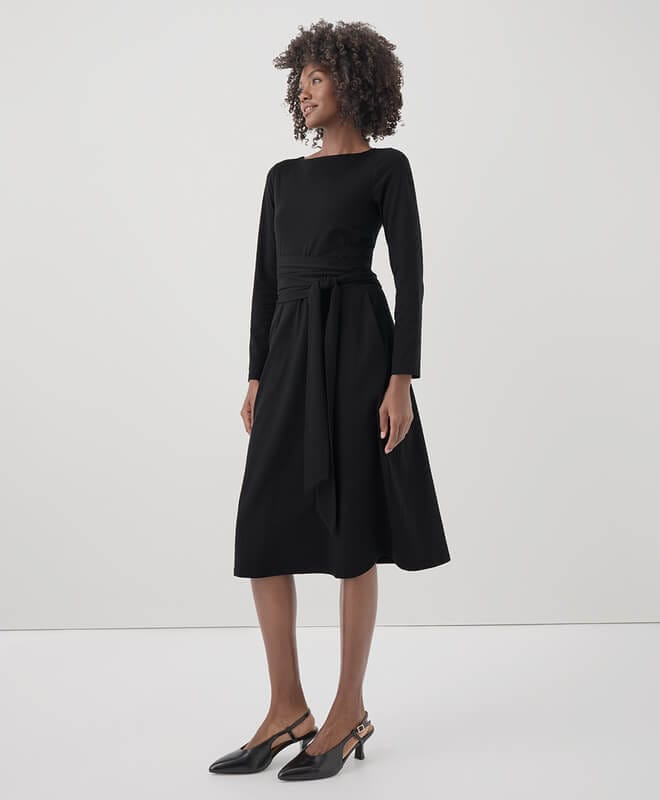 A woman standing against a white background wearing a wrap black dress.