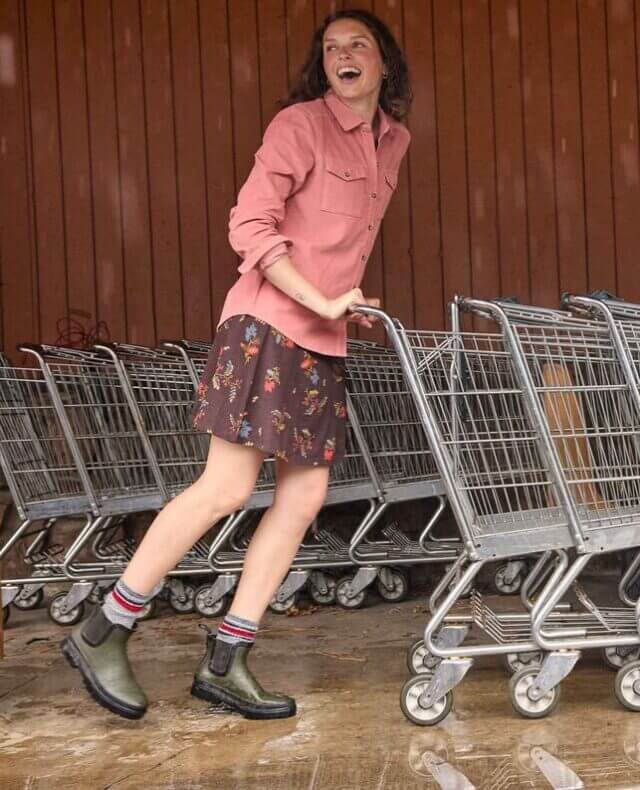 A smiling woman pushing trolleys wearing a pink shirt and a black printed skirt.