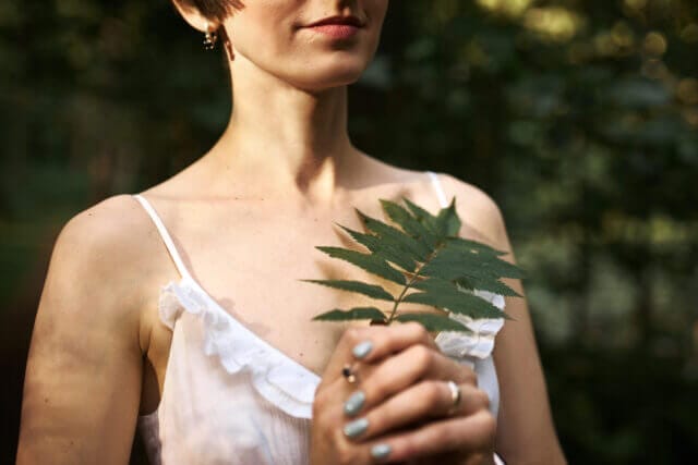 Unrecognizable young woman with short hairstyle and pale skin, holding green fern leaf.
