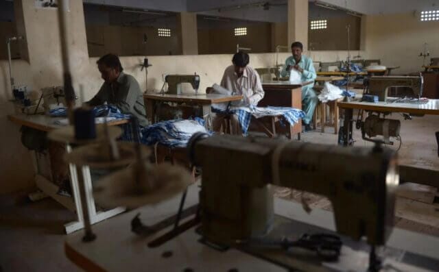 Garment workers sewing clothes for fast fashion clients in a dimly lit factory, highlighting the challenging working conditions they endure.