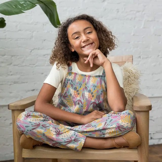 Smiling girl sitting cross-legged in a wooden chair, wearing colorful organic cotton overalls with a botanical print.