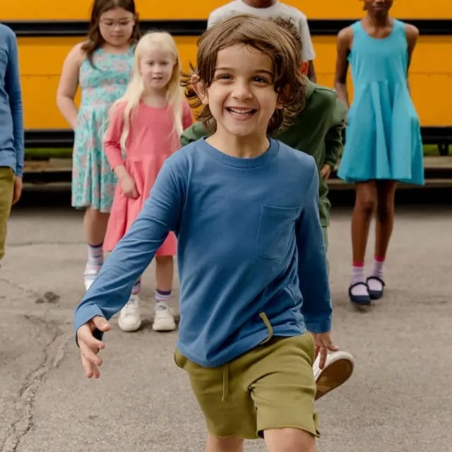 Young boy smiling and running toward the camera with other children walking behind him near a yellow school bus.