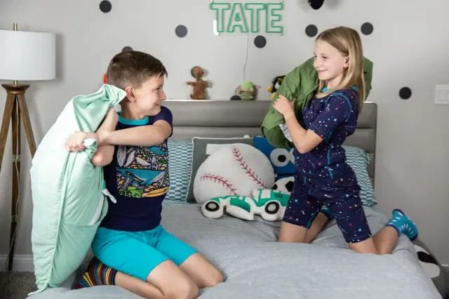 Two kids laughing and having a pillow fight on a bed, wearing bright organic cotton pajamas.