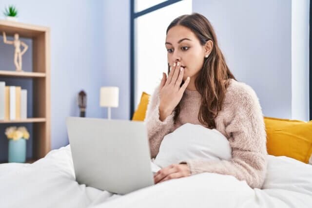 shocked woman reads article in bed on her laptop. Meant to represent the shocking unsustainability of Edikted.
